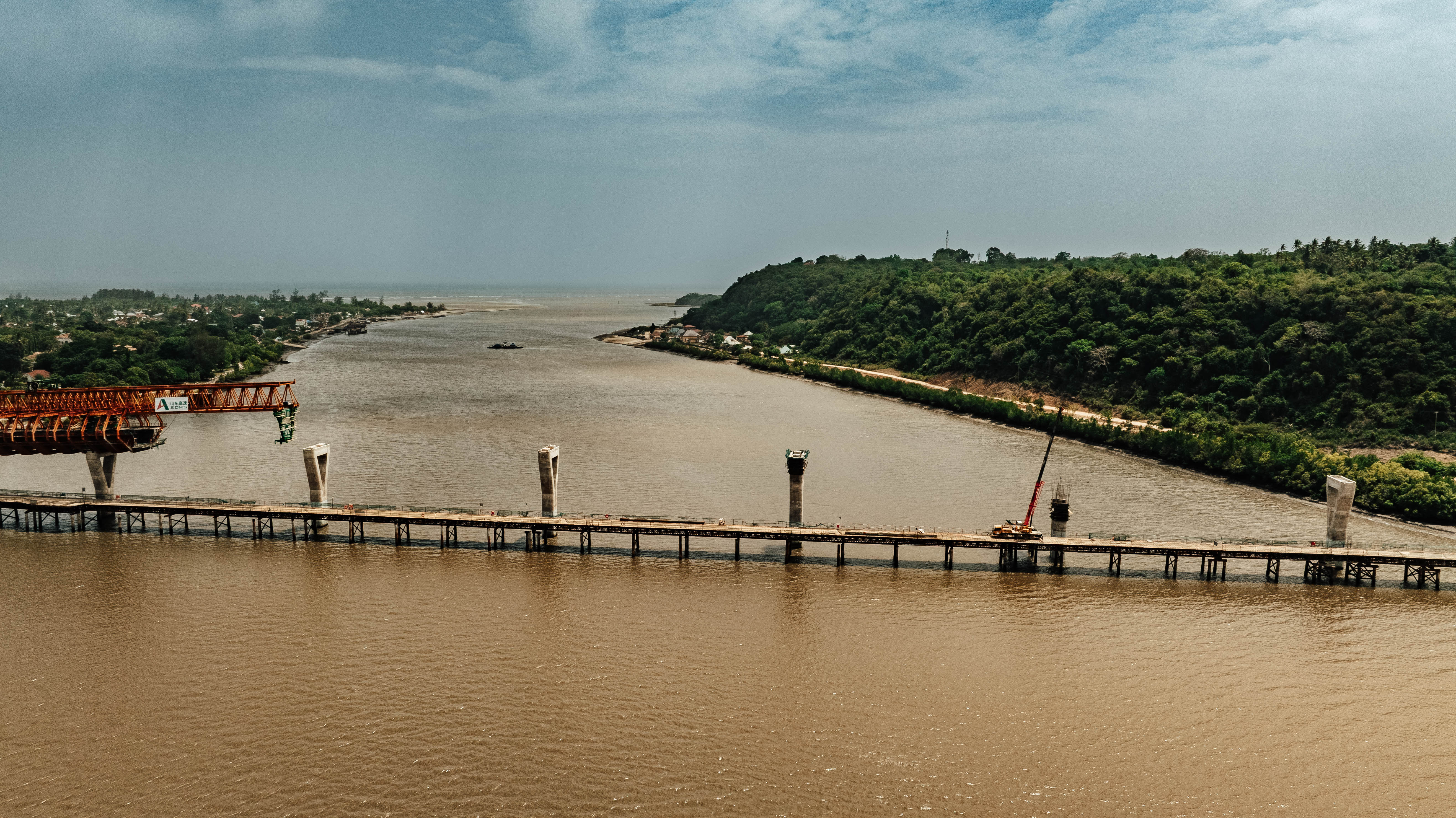 A partially constructed bridge spans a wide river, surrounded by greenery and distant infrastructure.