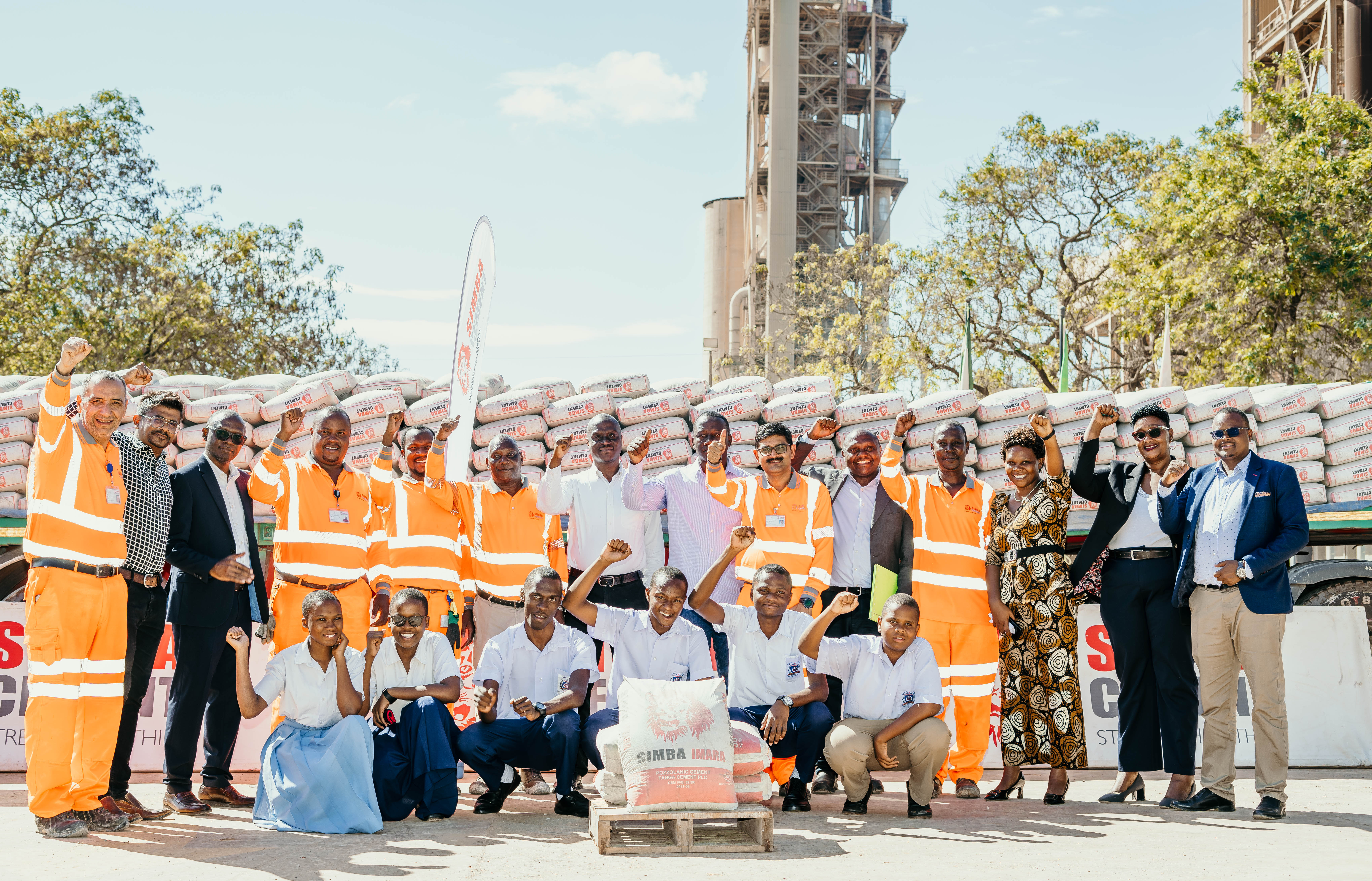 A group of people, including workers in orange safety vests, pose in front of stacked cement bags at an industrial site.