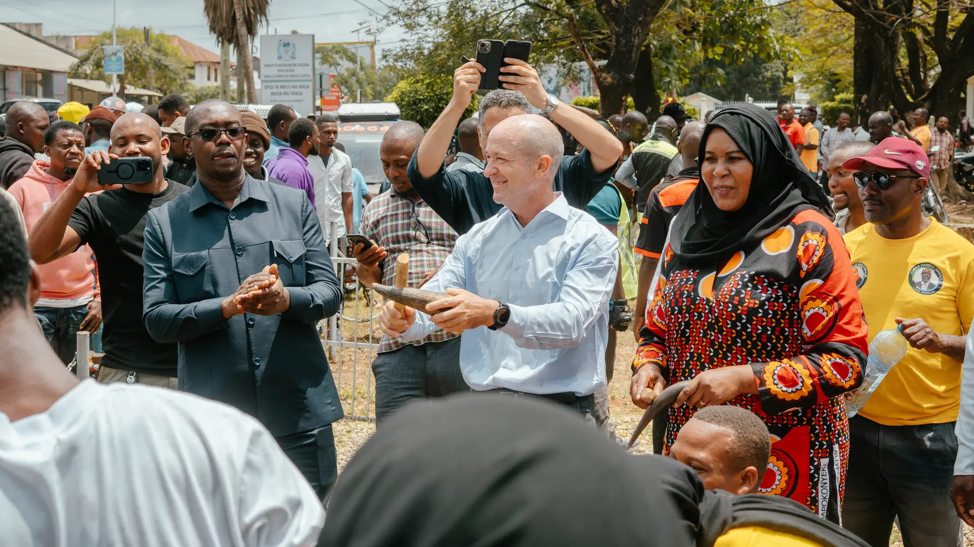 A group of people gathered outdoors, with some holding tools and observing an activity, possibly related to a community or construction event.