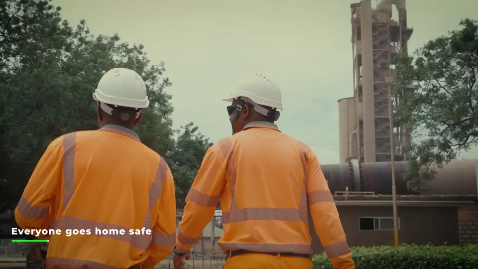 Two workers in safety gear walk near an industrial site with a large building visible in the background.